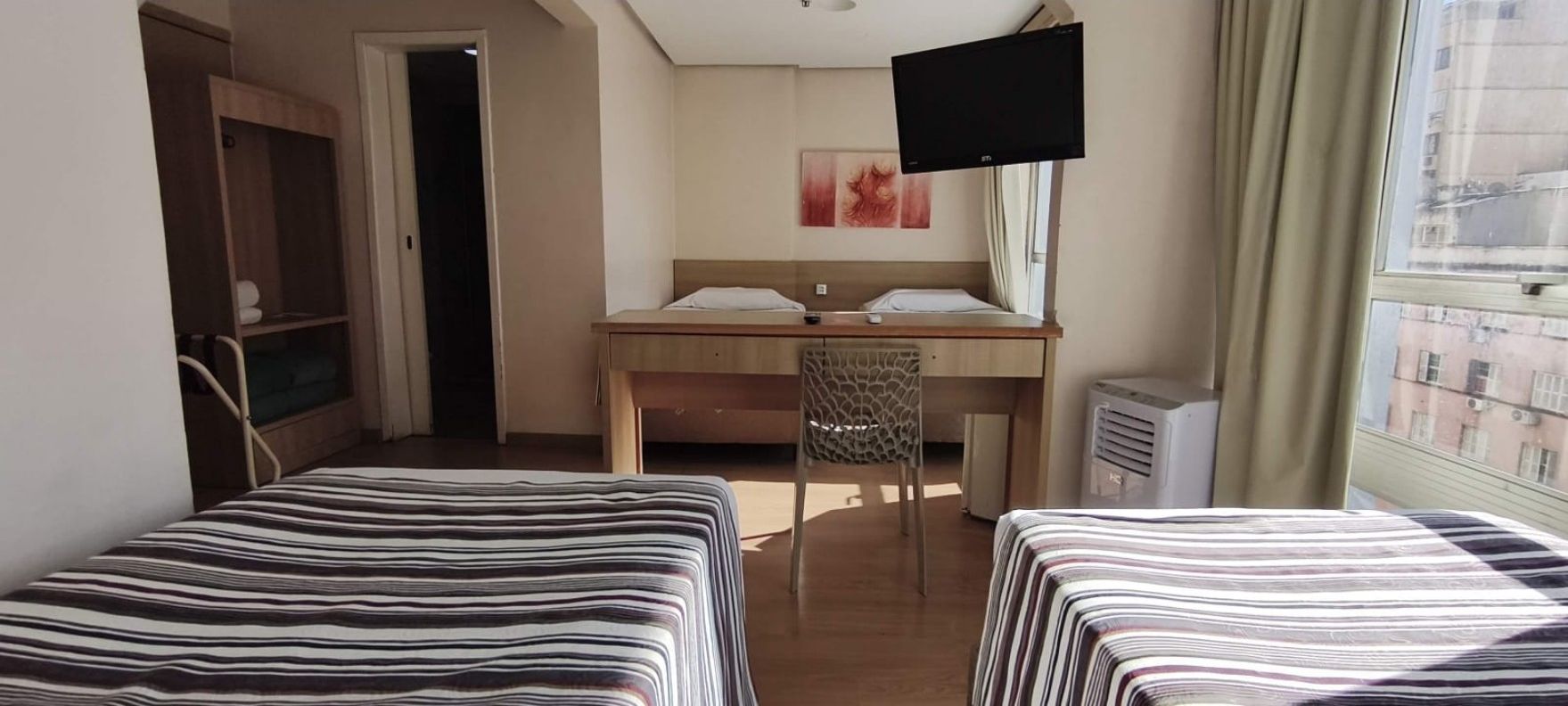 Ein Hotelzimmer mit zwei Betten, einem Schreibtisch, einem Stuhl, einem Fernseher und einem Fenster mit Blick auf Gebäude.