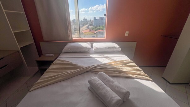 A hotel room with a bed, pillows, a bedside table, a phone, and a window with a city view. - Hotel Boreas (Fortaleza)