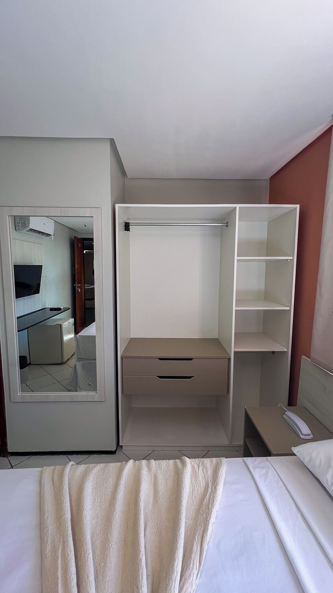 Habitación de hotel compacta con cama, escritorio pequeño, espejo y armario con estantes.