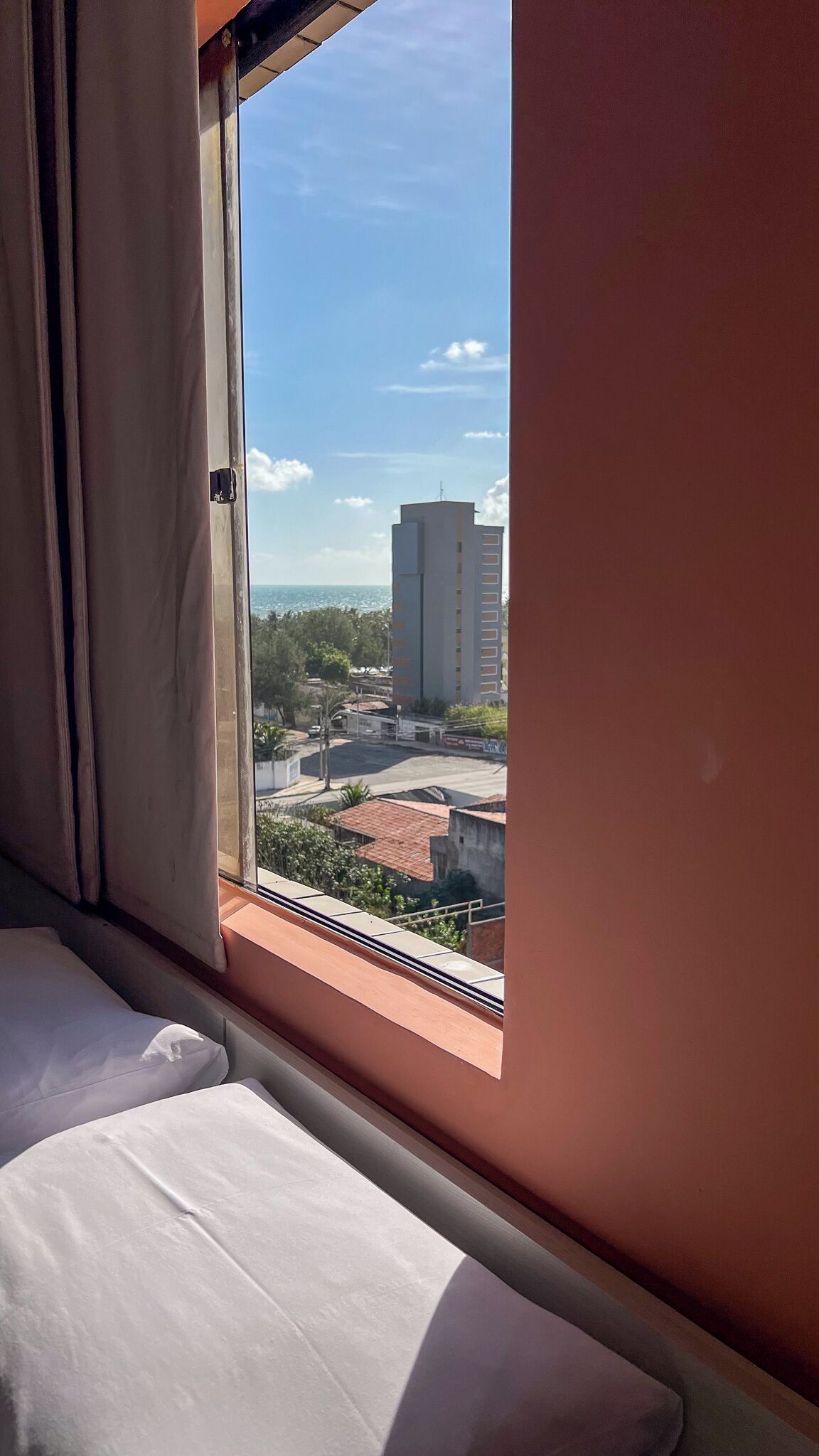 A hotel room with a bed, a window with curtains, and a view of a cityscape including buildings and greenery.