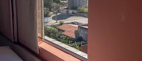 A hotel room with a bed, a window with curtains, and a view of a cityscape including buildings and greenery.