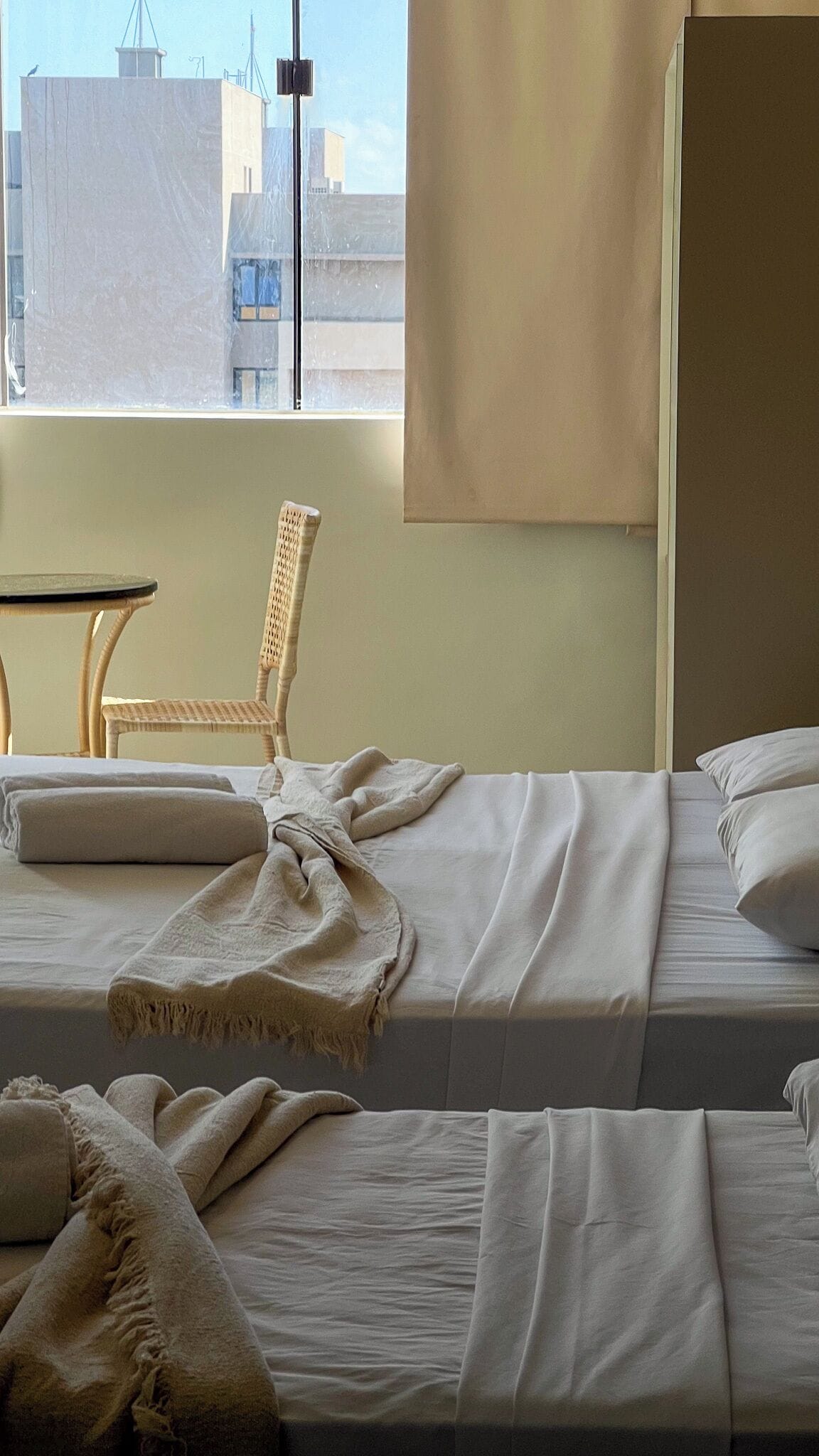Habitación de hotel con dos camas, una silla de mimbre, una mesa y un ventanal con vistas a edificios.