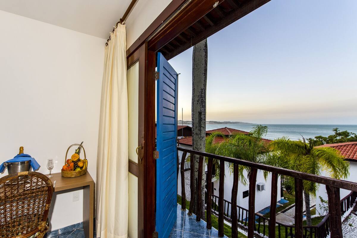 A balcony with a view of the sea, a wicker chair, a table with a bottle and a fruit basket, and a blue door.