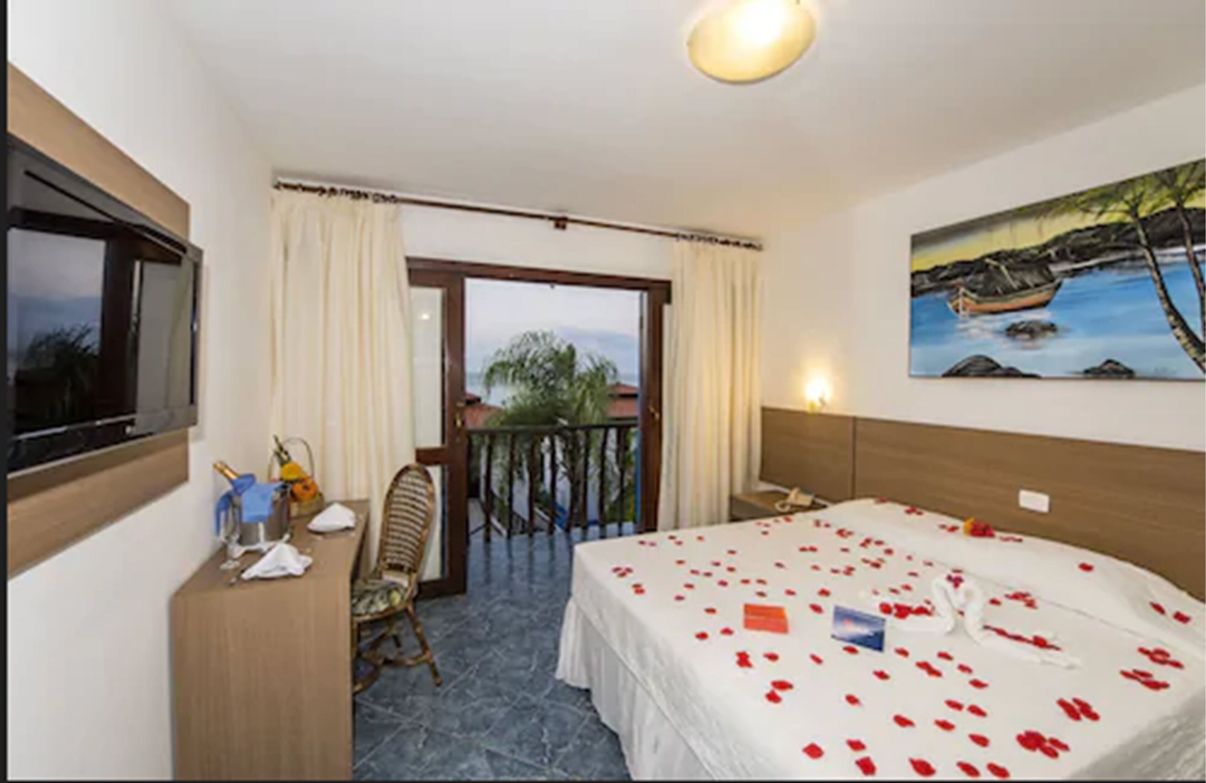 Habitación de hotel con una cama cubierta de pétalos de rosa, un cuadro de un barco en la pared y un balcón con cortinas.