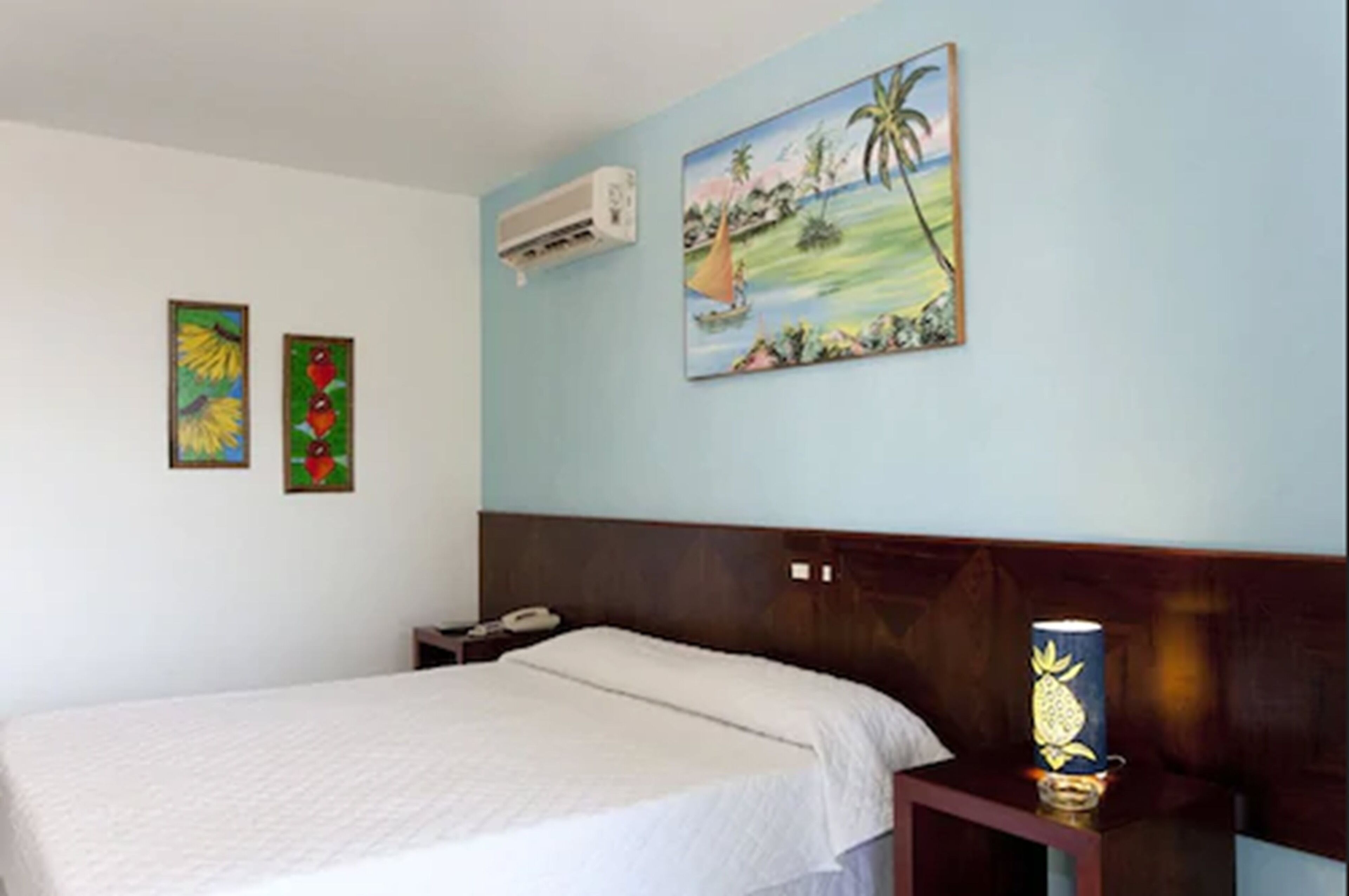 A hotel room with a bed, a nightstand with a lamp, and two paintings on the wall.
