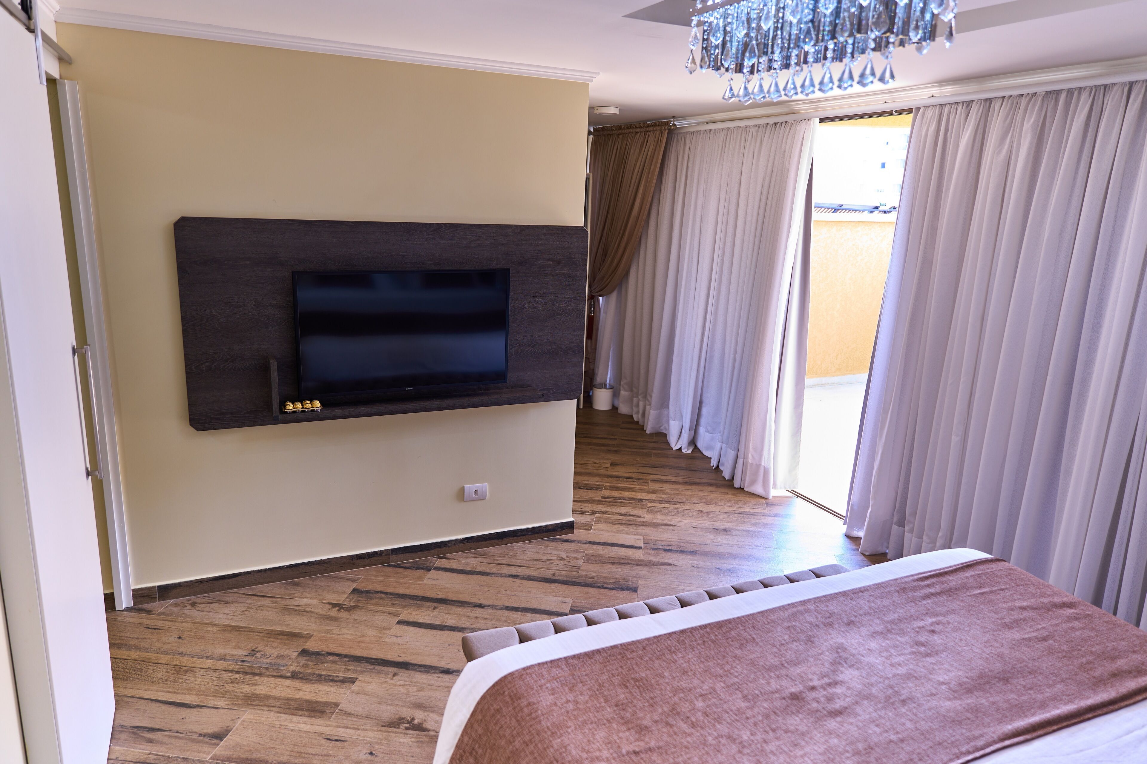 A hotel room with a large bed, a mounted TV, wooden flooring, and a view of the outside through the curtains.