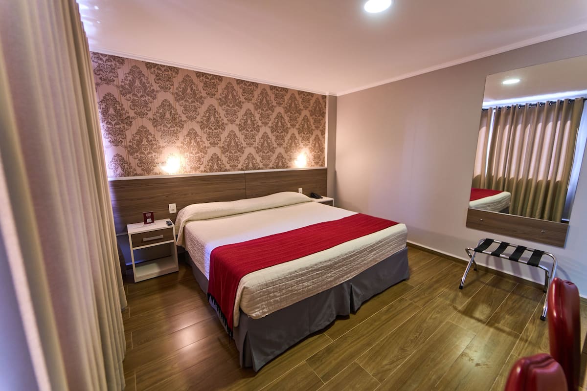 A hotel room with a bed, nightstand, mirror, and wooden flooring.