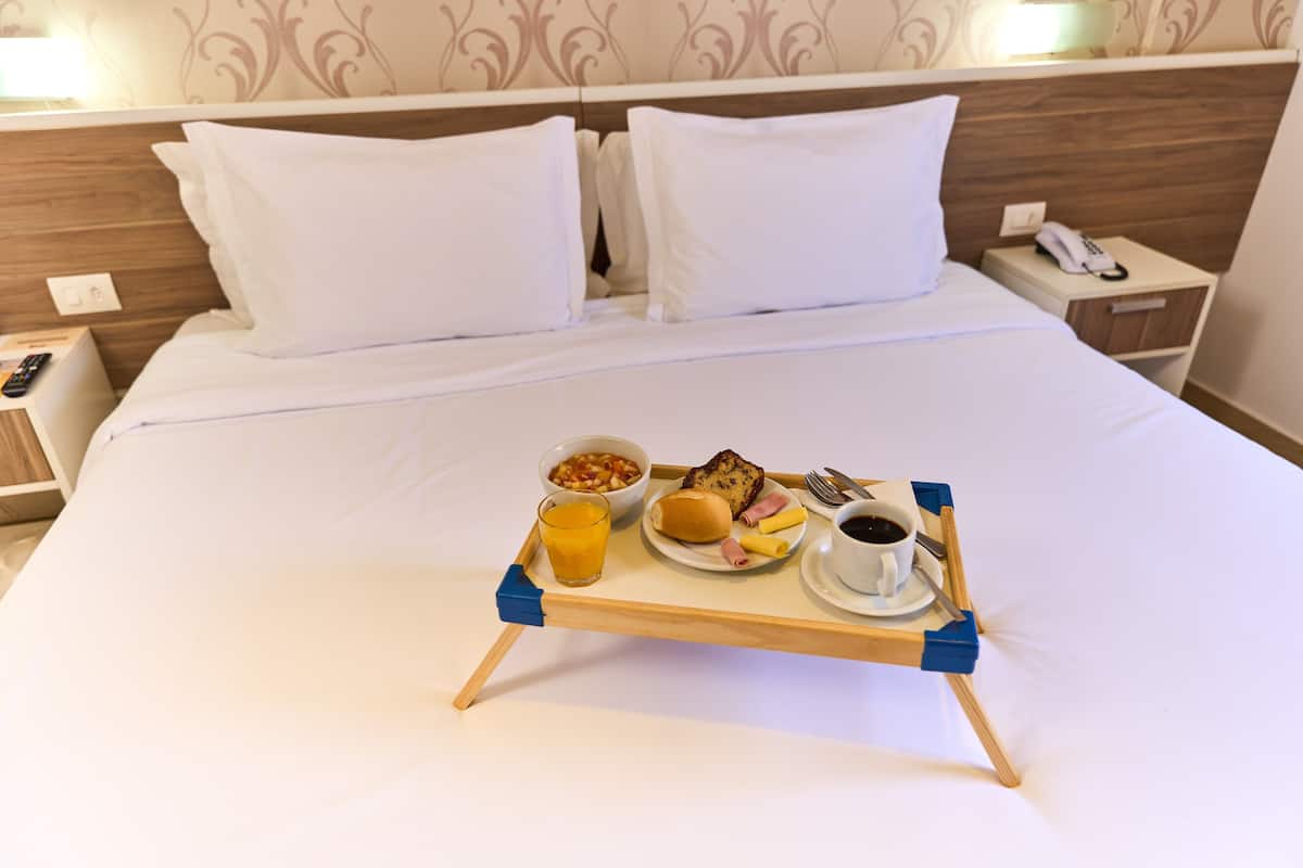 A neatly made bed with white linens, a bedside table with a phone, and a tray with breakfast items including toast, eggs, and coffee.