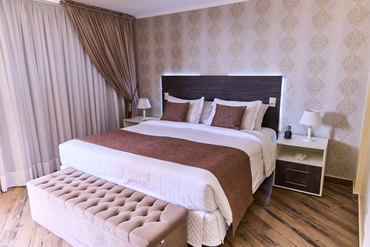 A large bed with a wooden headboard, a bedside table with a lamp, and a bench at the foot of the bed.