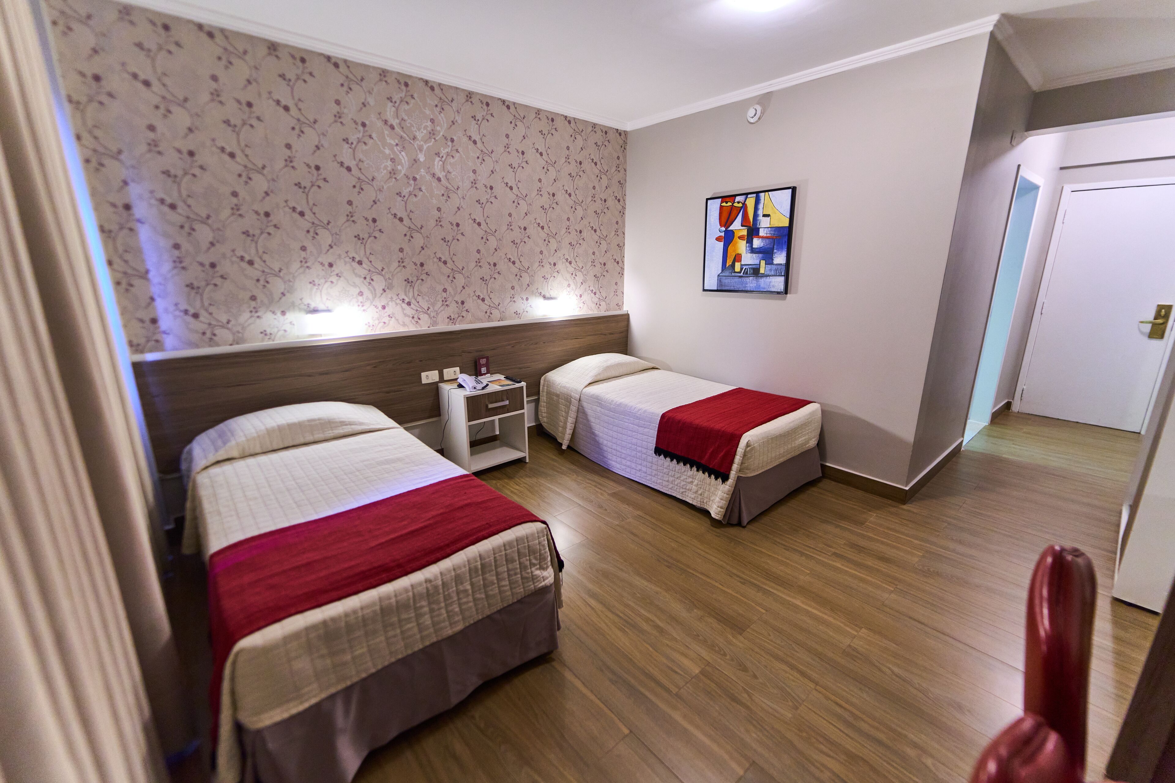 Quarto de hotel com duas camas, uma mesa de cabeceira, um quadro na parede e uma porta à direita.