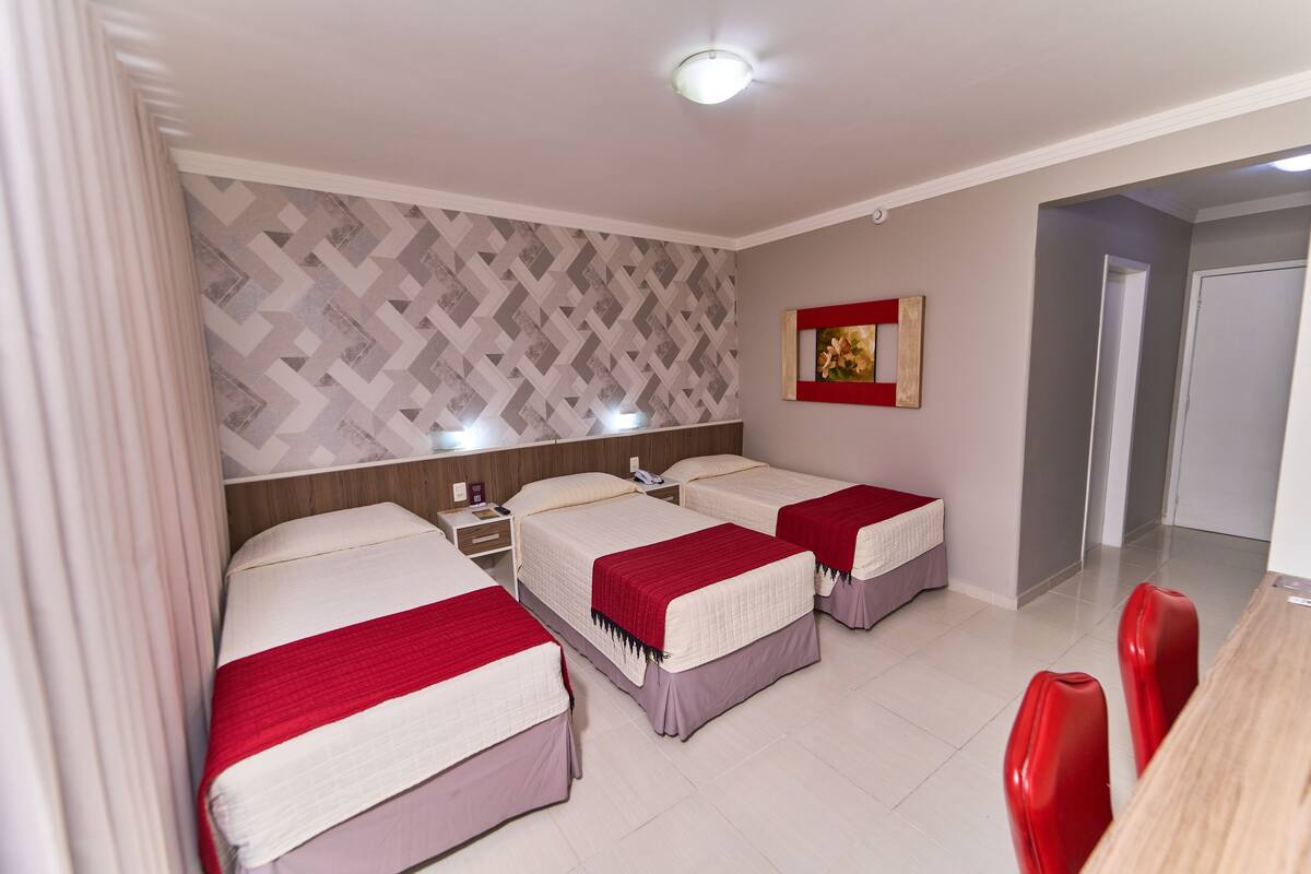 A hotel room with three beds, a dining table with red chairs, a patterned wall, and a painting on the wall.