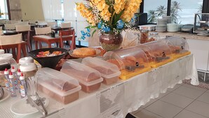 A buffet setup with various food items, a vase of flowers, and a view of palm trees through the window. - Golden Beach Hotel (Fortaleza)