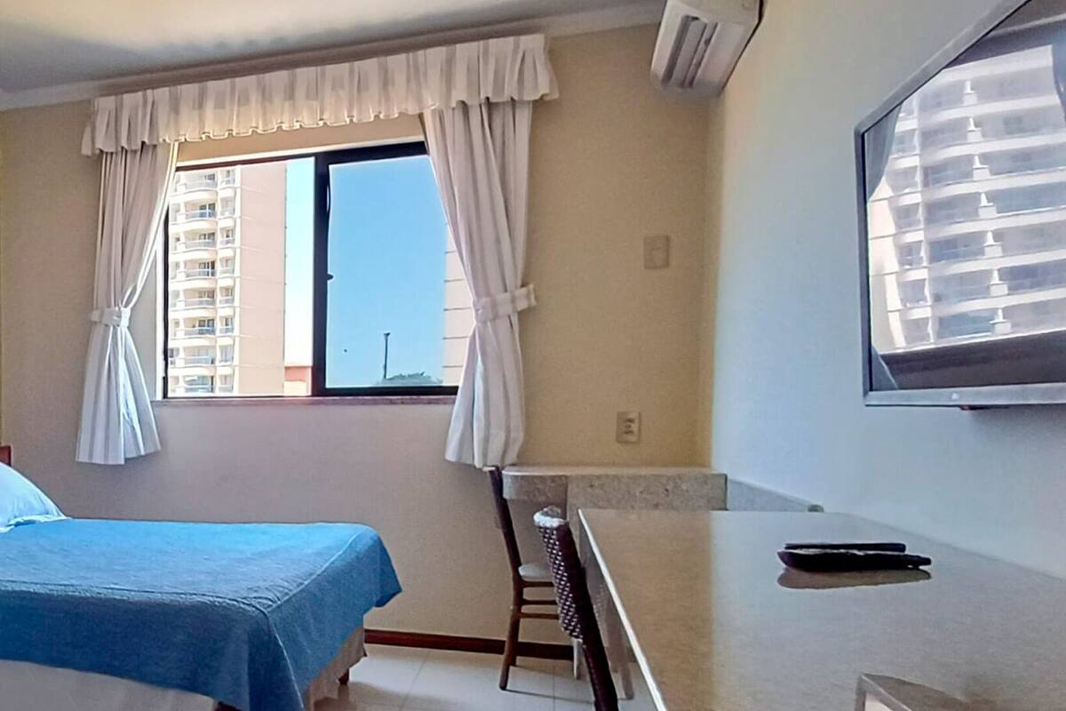 Hotellihuone, jossa on sänky, pieni työpöytä, tuoli, ikkuna verhoilla ja ilmastointilaite.