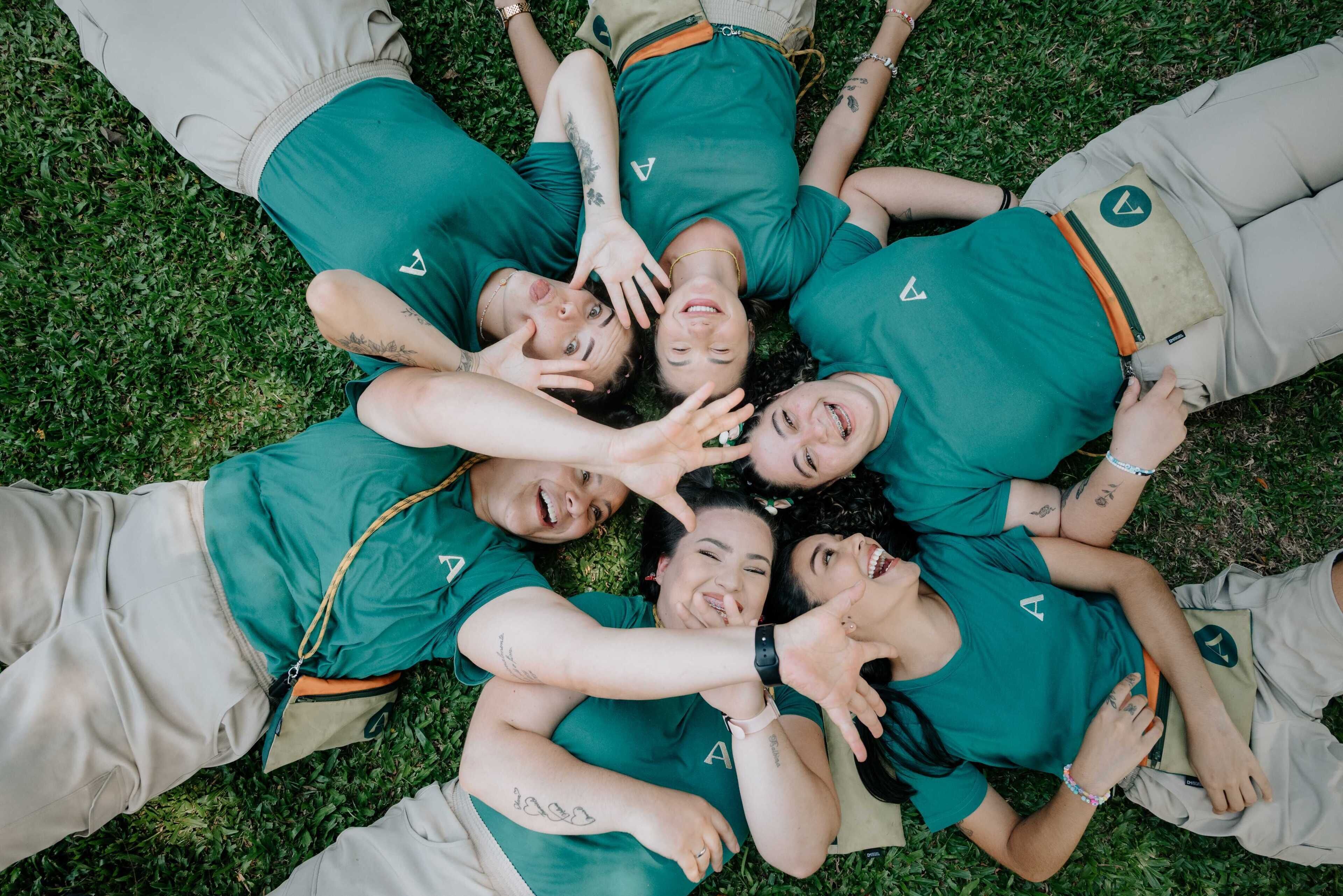 A group of people lying on grass in matching green shirts and beige pants.