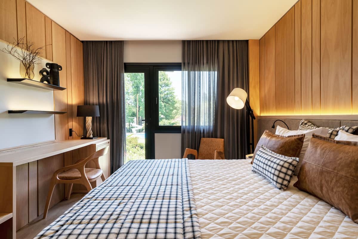 A modern hotel room with a wooden panel wall, a desk with a chair, a bed with a quilted cover, and a view of the outdoors through a large window.