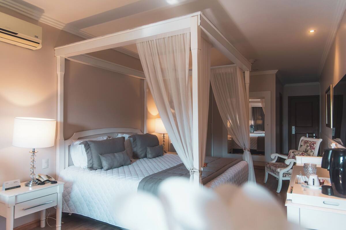 A four-poster canopy bed with a bedside table and lamp, a chair, and a small table with a coffee maker.