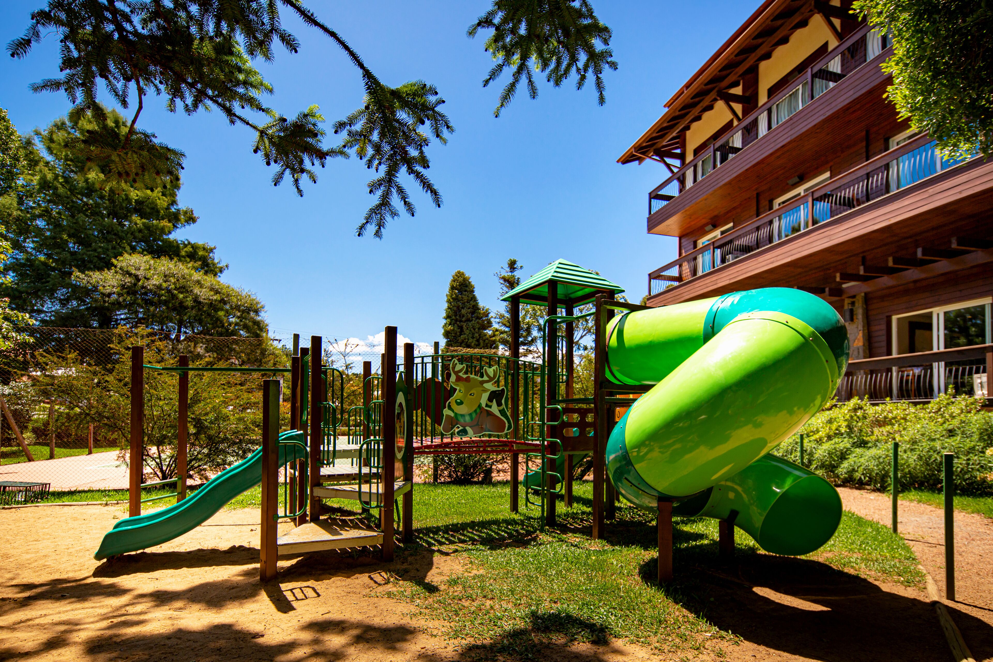 Un parque infantil con tobogán y estructuras para escalar, rodeado de árboles y un edificio con balcones.