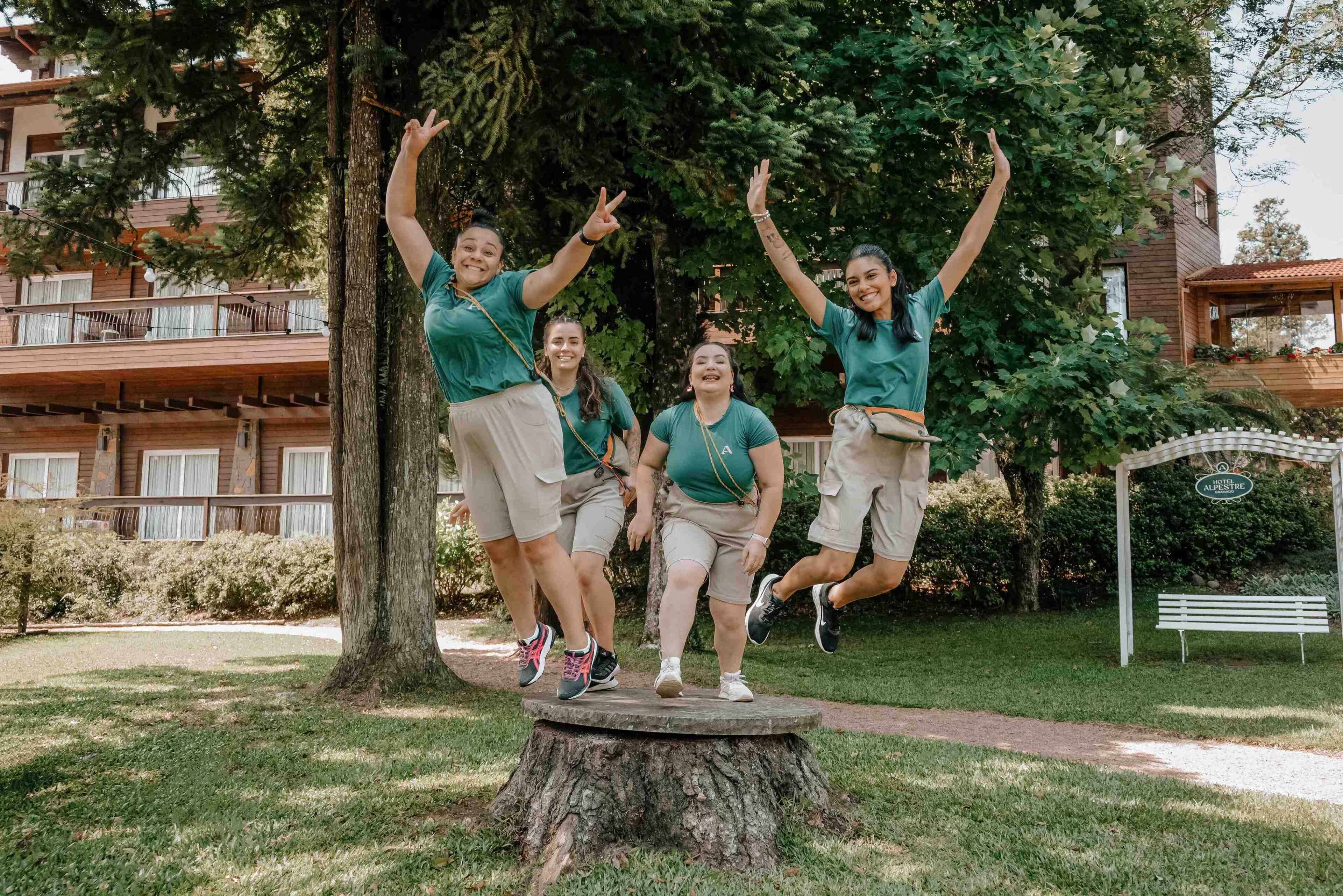 A group of people in green shirts and beige shorts jumping on a tree stump in a park with a building in the background.