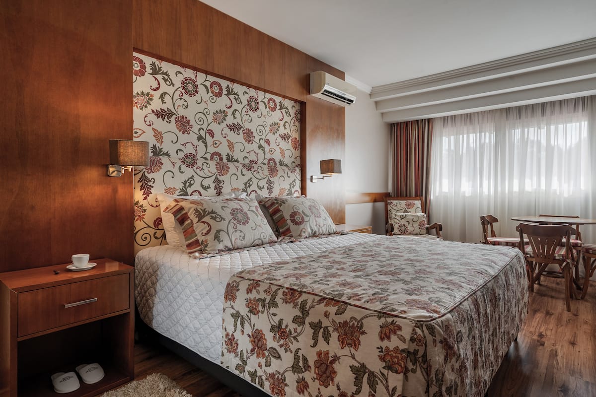 A hotel room with a wooden headboard, floral-patterned bedspread, a bedside table with a cup and slippers, and a small table with chairs.
