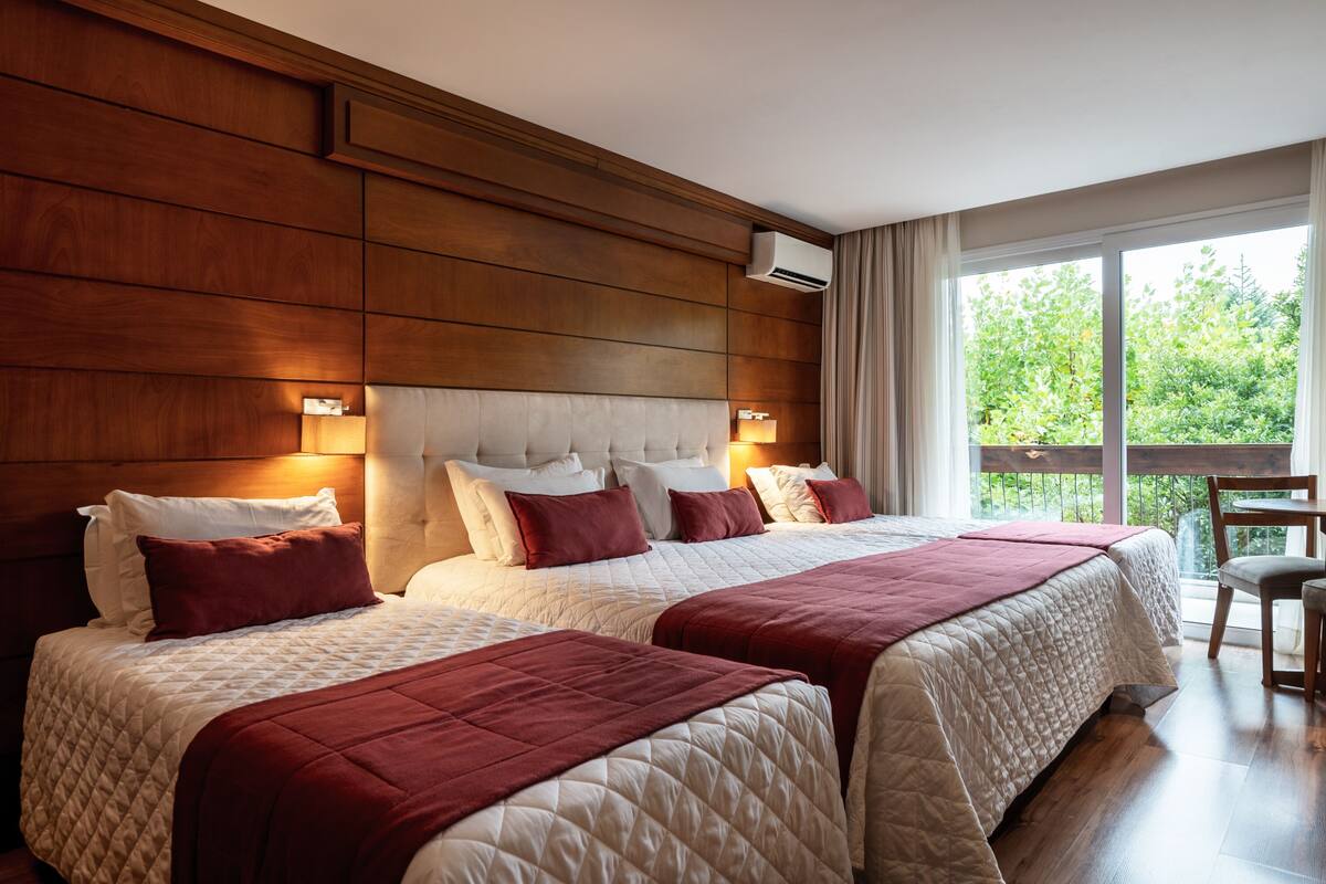 A hotel room with two beds, wooden paneling, a large window with a view of greenery, and a chair by the window.