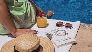 Una persona sentada junto a una piscina, con sombrero de paja, gafas de sol, una bebida con fresa y un libro titulado 'GPEA McKEDOWN'.