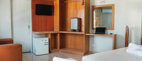 A hotel room with a bed, a small refrigerator, a wooden wardrobe, a desk with a laptop, and a mirror.