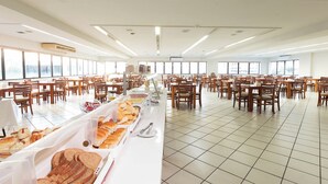 Un grand espace dédié à la restauration, avec de nombreuses tables et chaises, un buffet proposant du pain et des viennoiseries, et de grandes fenêtres laissant entrer la lumière naturelle.
