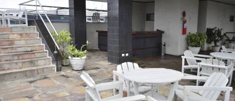An outdoor patio with white chairs and tables, stone flooring, a fire extinguisher, and potted plants.