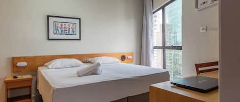 A hotel room with a bed, a desk with a laptop, a nightstand, a framed picture on the wall, and a window with a view of buildings.