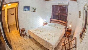 Un dormitorio con cama, mesita de noche, taburete, ventana con contraventanas y puerta que da a otra habitación.