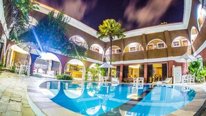Una piscina con diseño curvo, rodeada por un edificio de ladrillo con ventanas arqueadas y un patio cubierto.