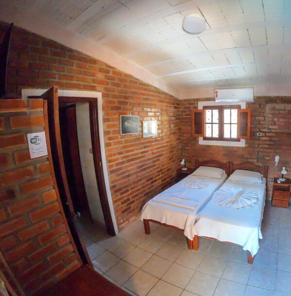 A room with a brick wall, a bed with a white bedspread, a bedside table, a nightstand, and a window with a view. - Real Pousada (Ipojuca)