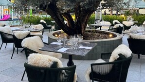 Un área de comedor al aire libre con mesas redondas, sillas negras con mantas de piel blanca y una chimenea central.