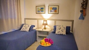 Habitación de hotel con dos camas, una mesita de noche, una lámpara y una cesta de frutas.