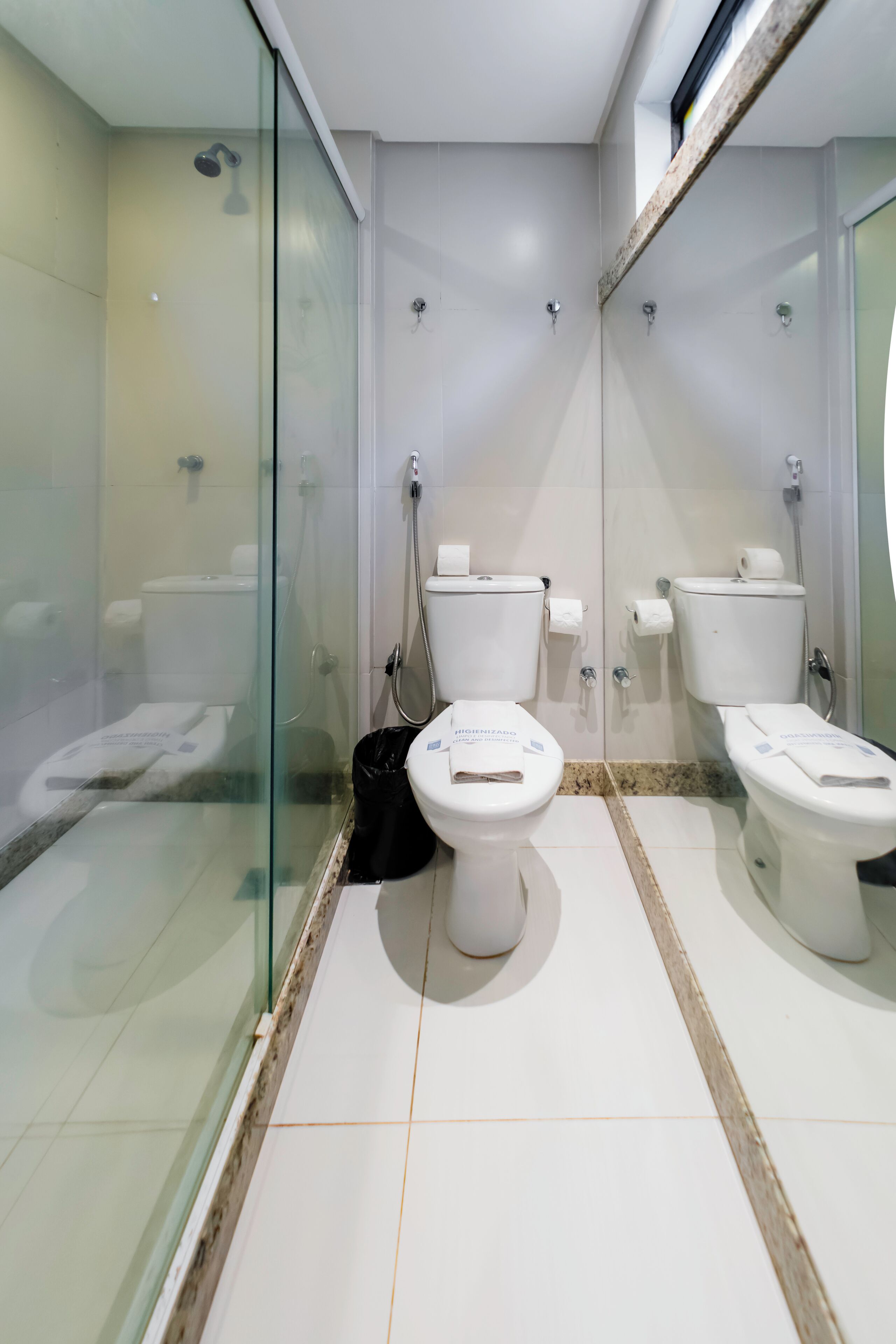 Un baño moderno con dos inodoros, una cabina de ducha de cristal y un cubo de basura negro.