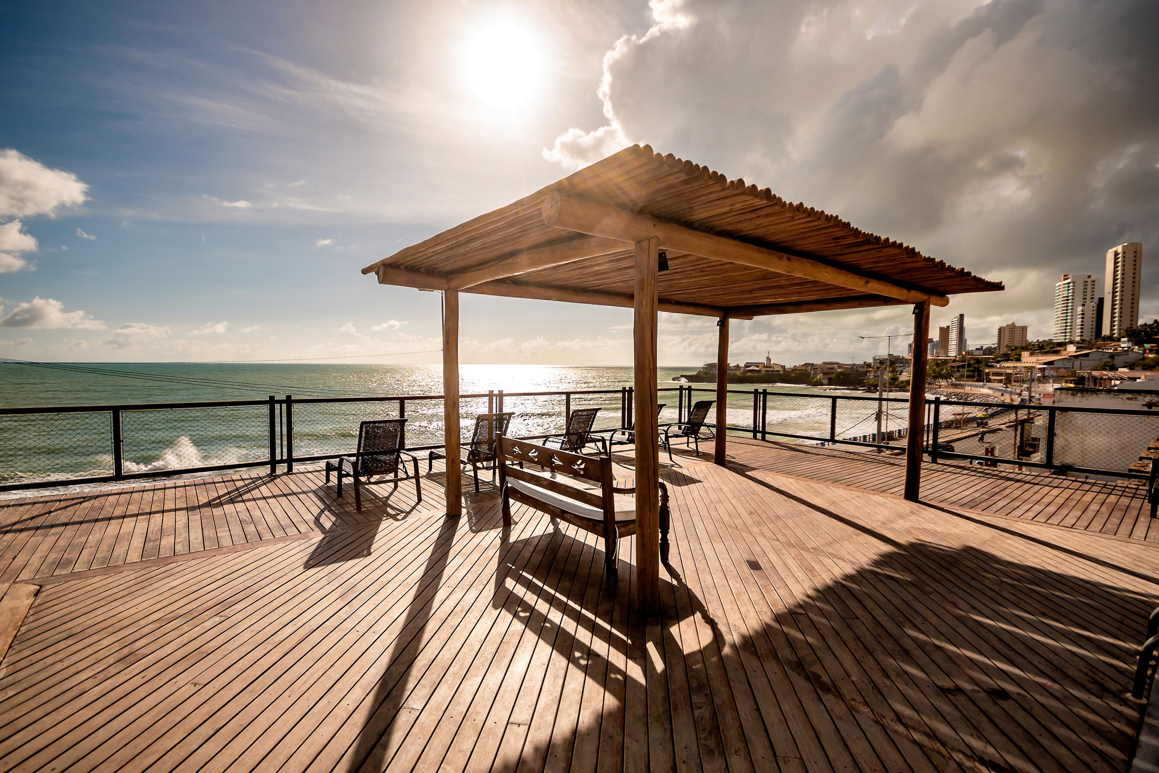 A wooden deck with a pergola, chairs, and a bench overlooking the ocean.