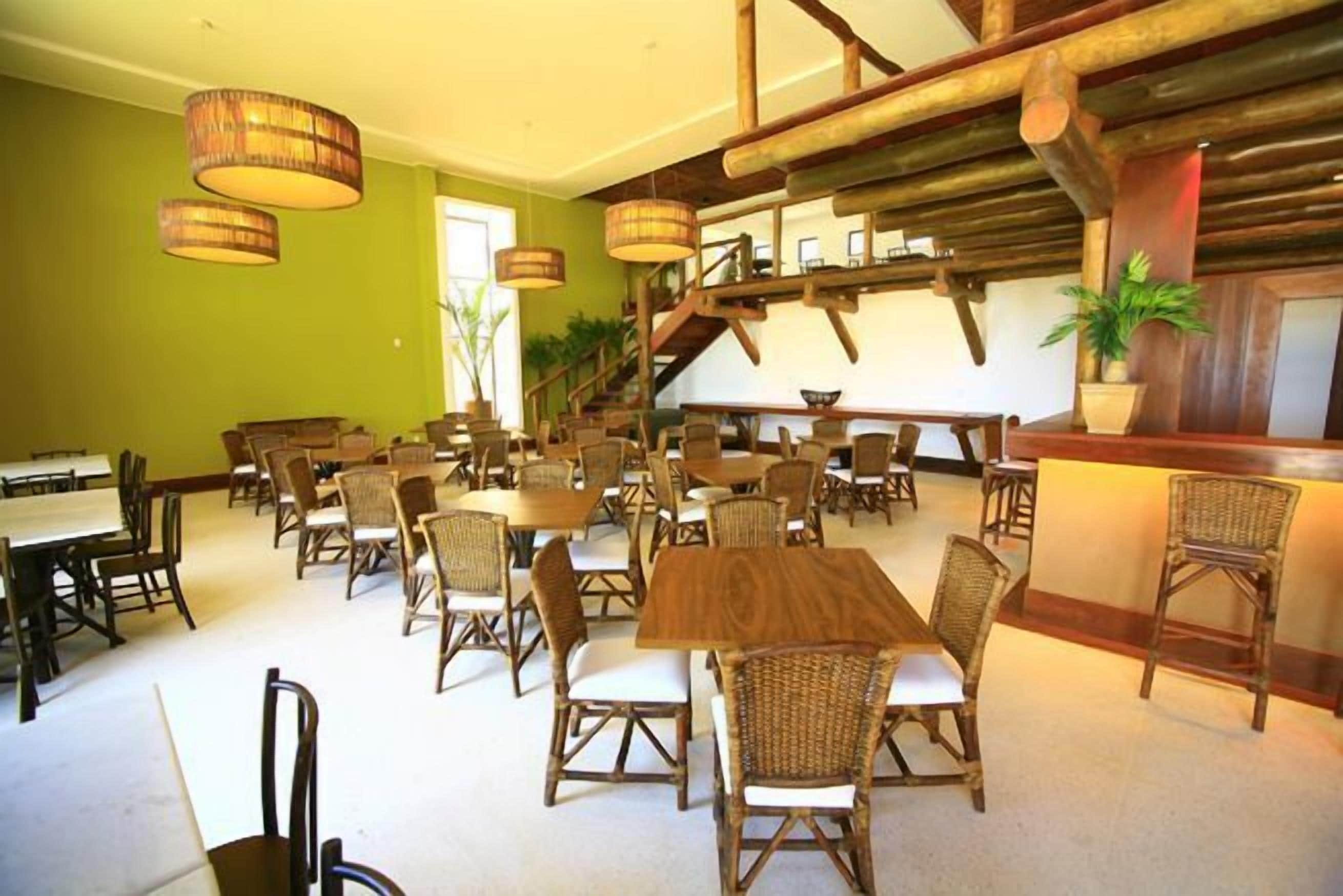 Uma sala de jantar espaçosa com várias mesas e cadeiras de vime, vigas de madeira no teto e uma escada que leva a um andar superior.