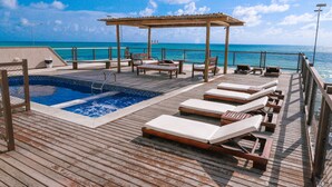Una terraza de madera con piscina, sillas de descanso y una pérgola con vistas al océano.