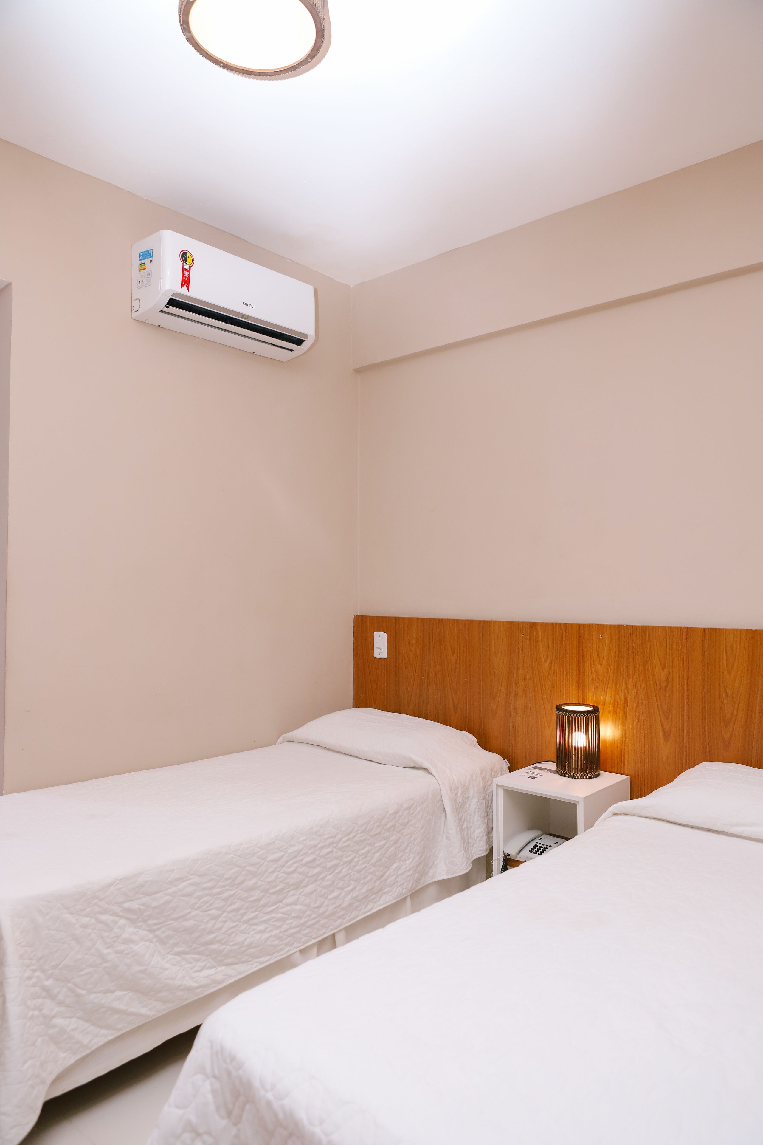 Habitación de hotel con dos camas individuales, cabecero de madera, unidad de aire acondicionado y lámpara de noche.