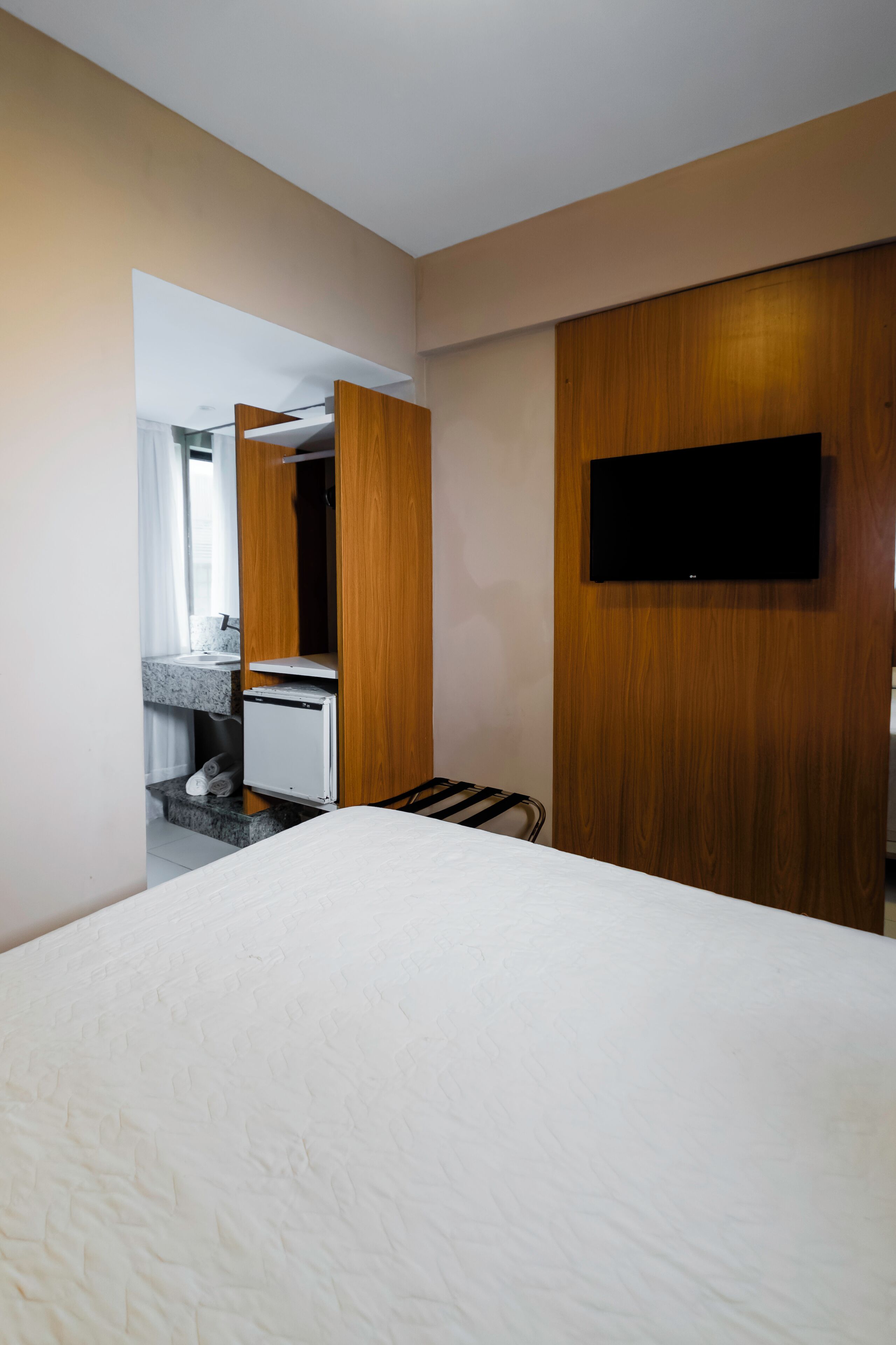 Habitación de hotel con cama, televisión montada en la pared, armario de madera y pequeña zona de kitchenette.