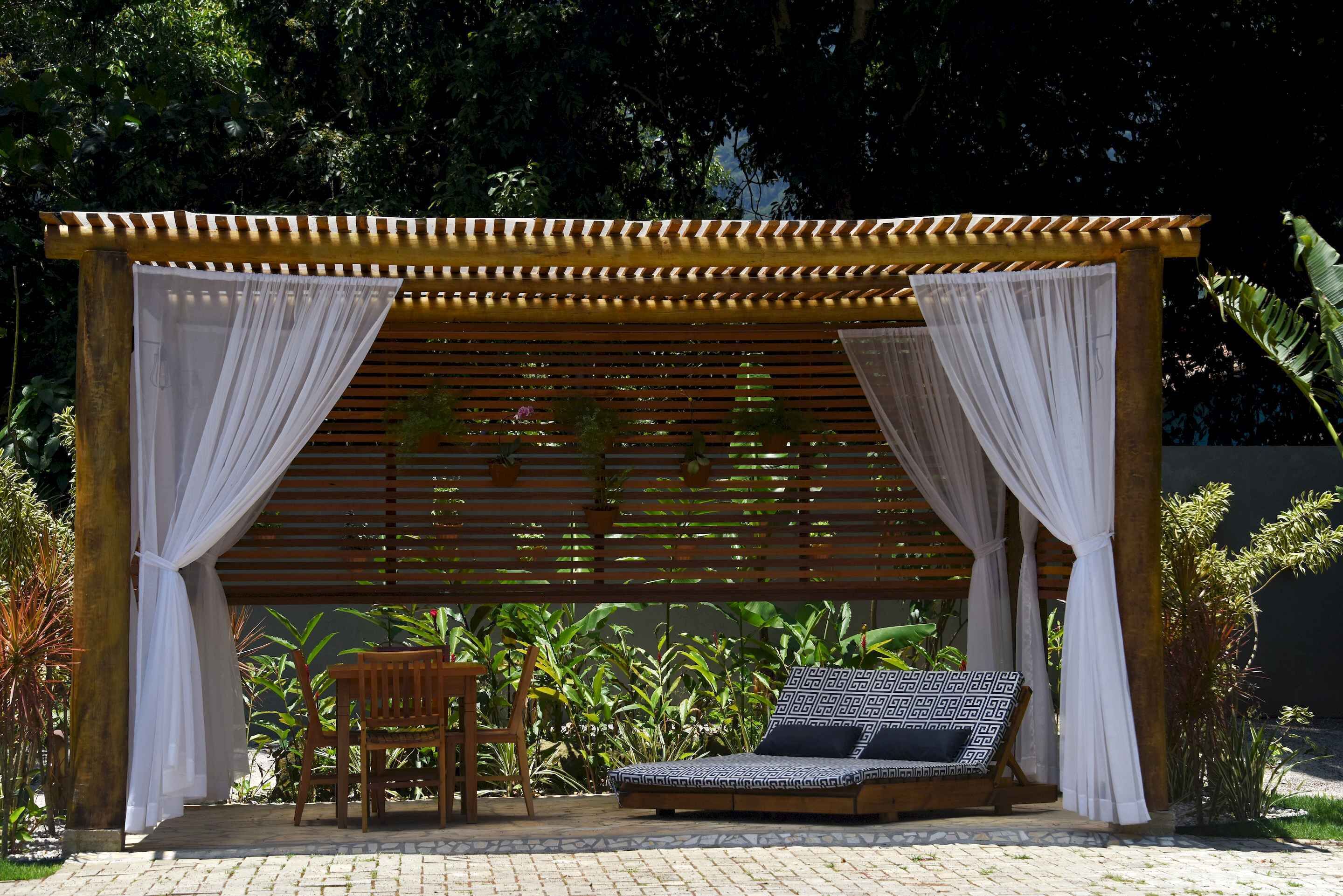 Uma pérgola de madeira com cortinas brancas, um banco de madeira e uma área de estar acolchoada com almofadas estampadas.