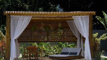 Uma pérgola de madeira com cortinas brancas, um banco de madeira e uma área de estar acolchoada com almofadas estampadas.