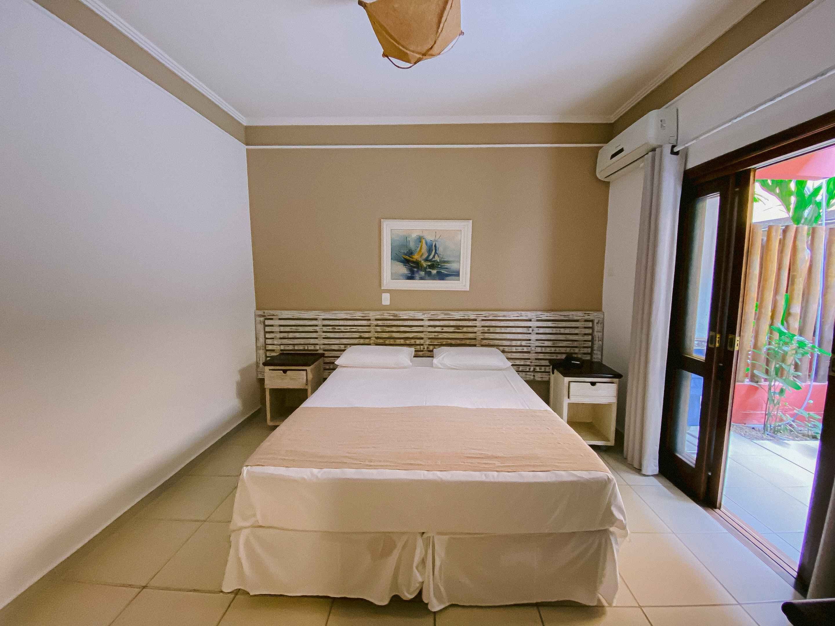 A hotel room with a large bed, a nightstand, a bedside table, a painting on the wall, and a sliding glass door to the outside.