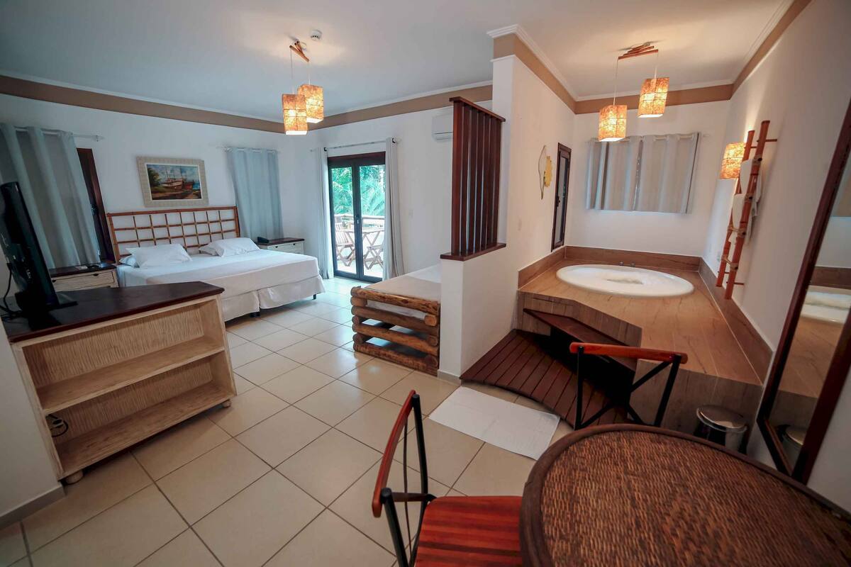 A hotel room with a bed, a wooden dresser, a bathroom with a bathtub, and a balcony with a view.