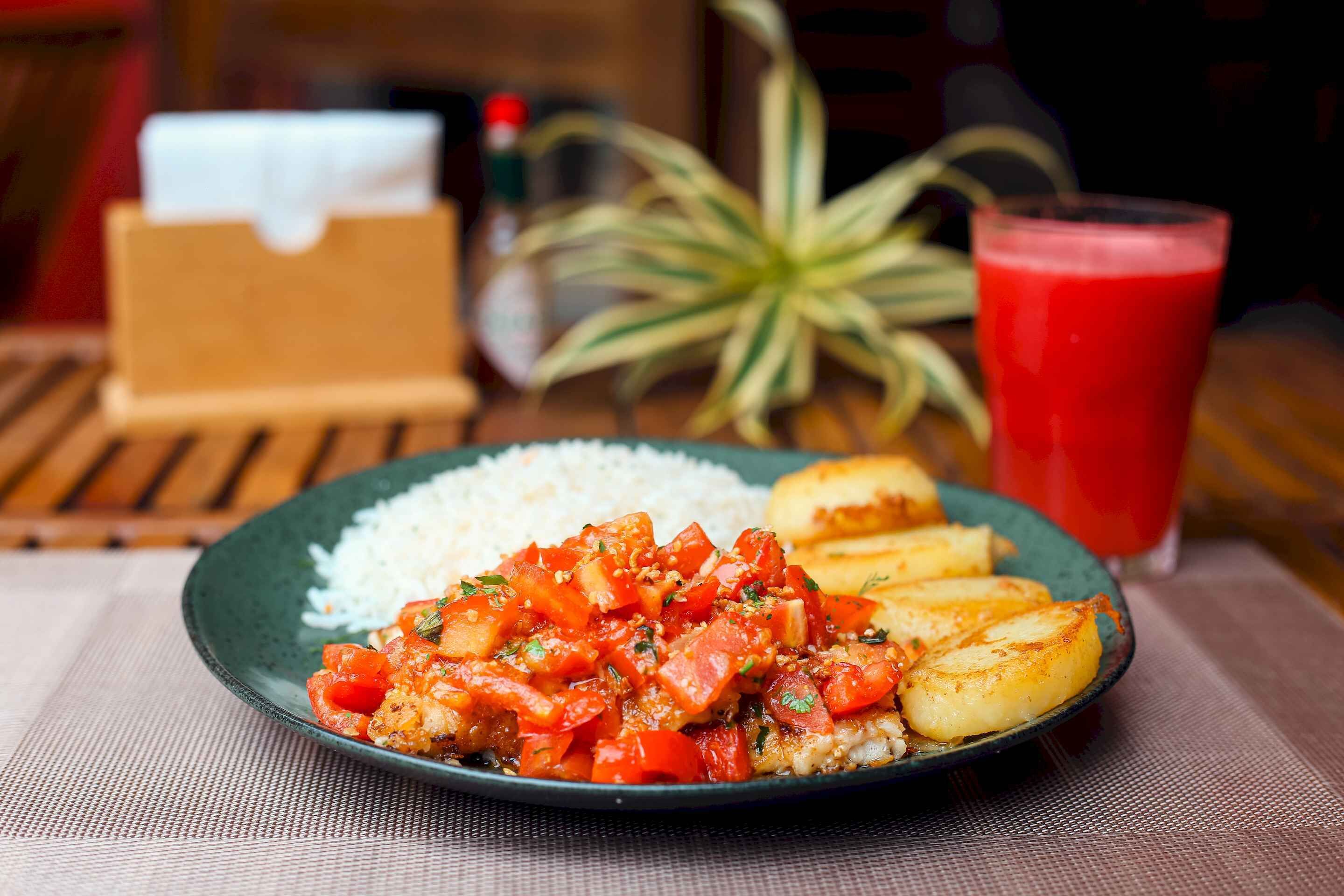 A plate of food with rice and a tomato-based dish, accompanied by fried plantains and a glass of red drink, on a table with a placemat.