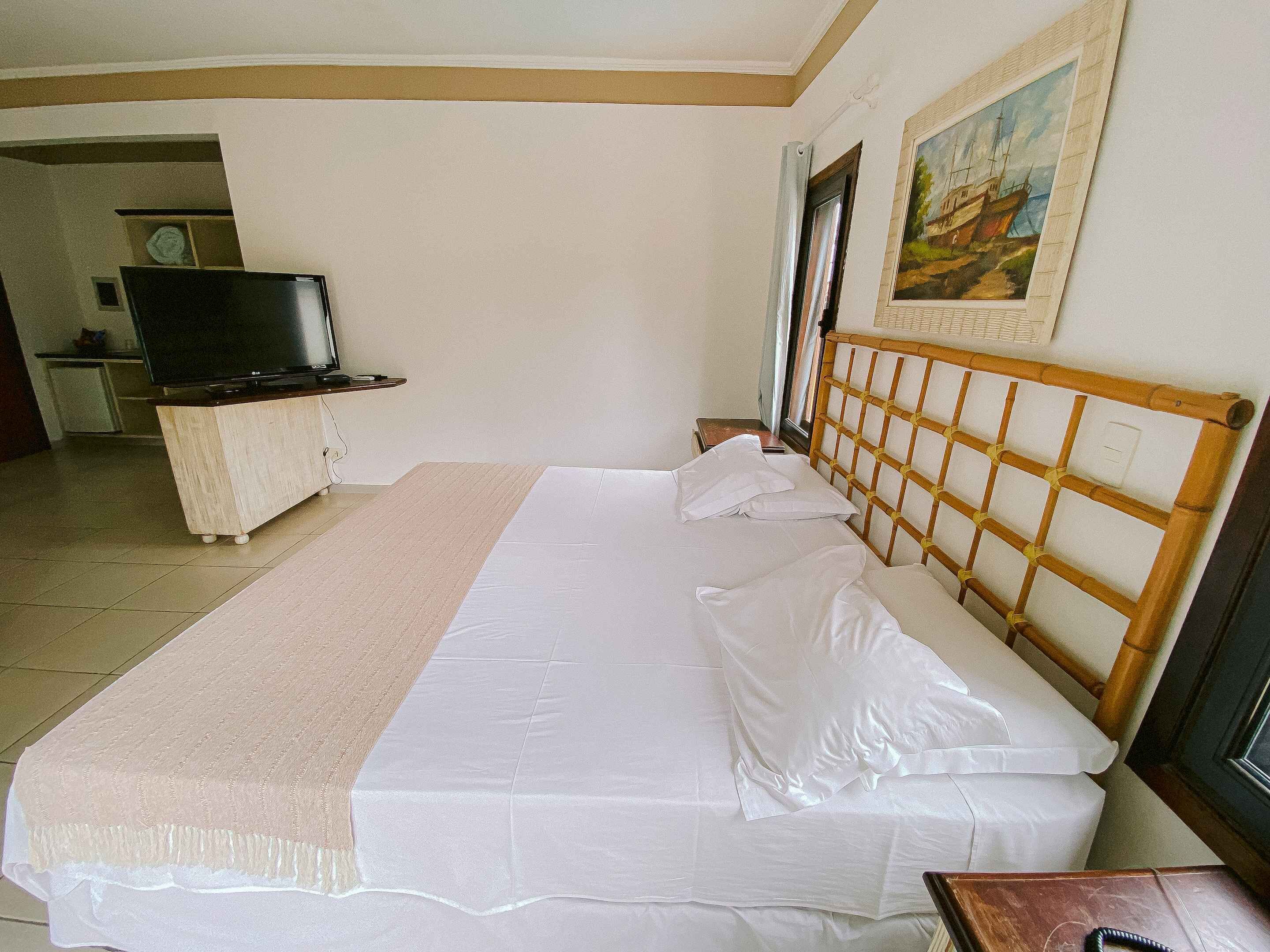 A neatly made bed with a wooden headboard, a bedside table, and a television in the background.