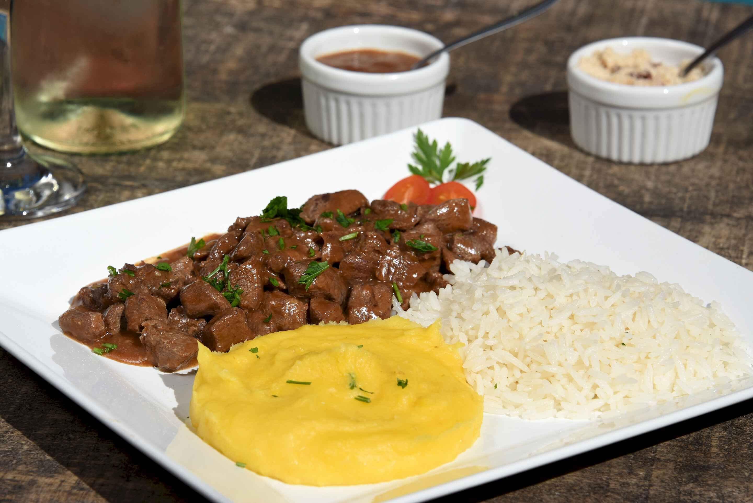 A plate with beef stew, rice, and a yellow side dish, possibly mashed potatoes, with a glass of white wine and small bowls of sauce in the background.