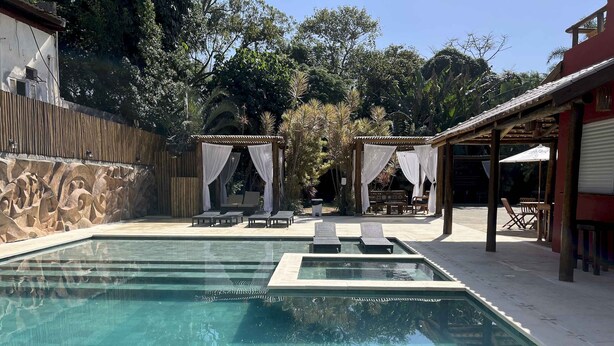 A swimming pool with lounge chairs, a covered seating area, and a stone wall. - Pousada Porto Mare (São Sebastião)