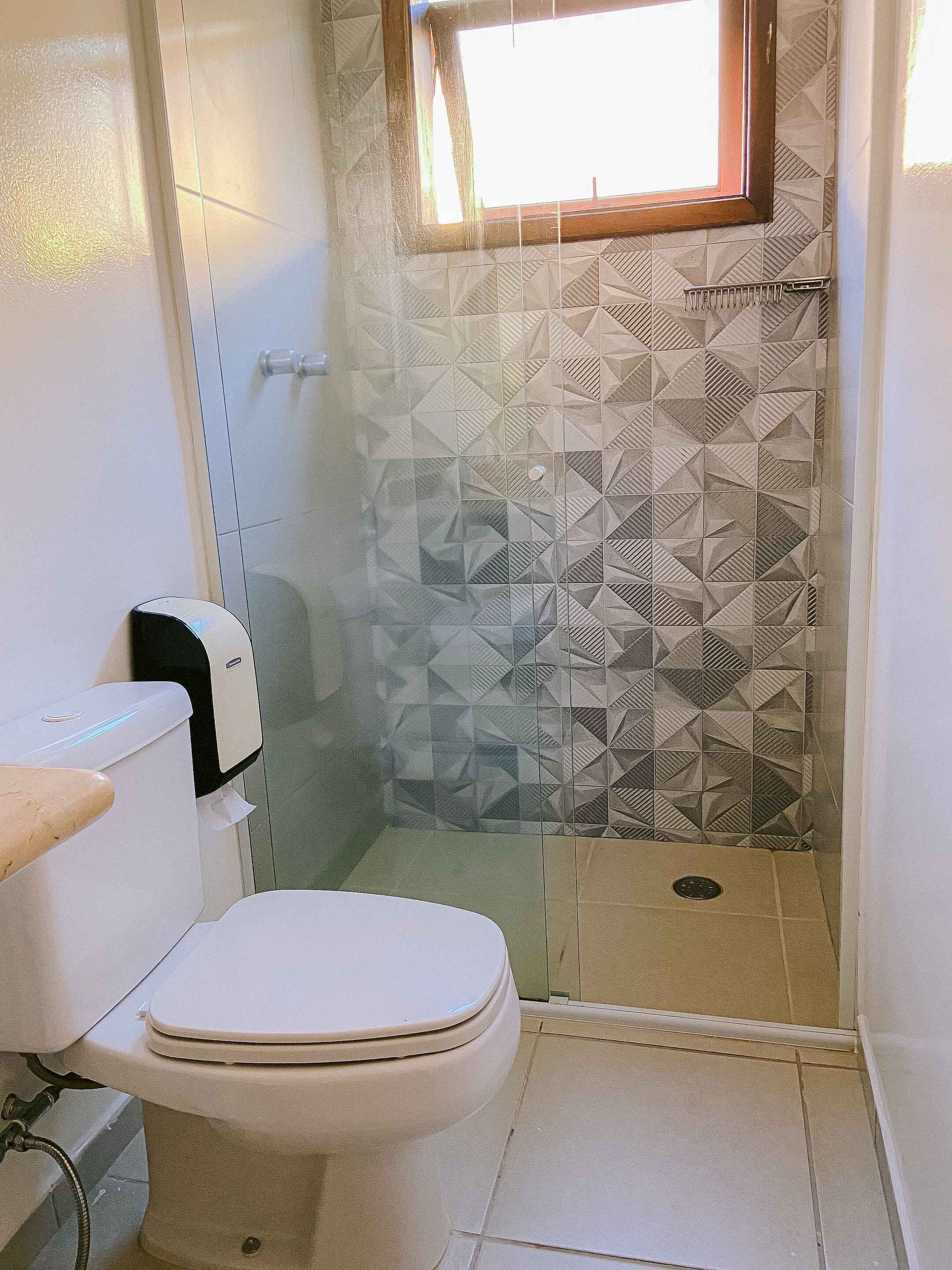 A bathroom with a toilet, a hand dryer, a shower with a geometric tile pattern, and a window.