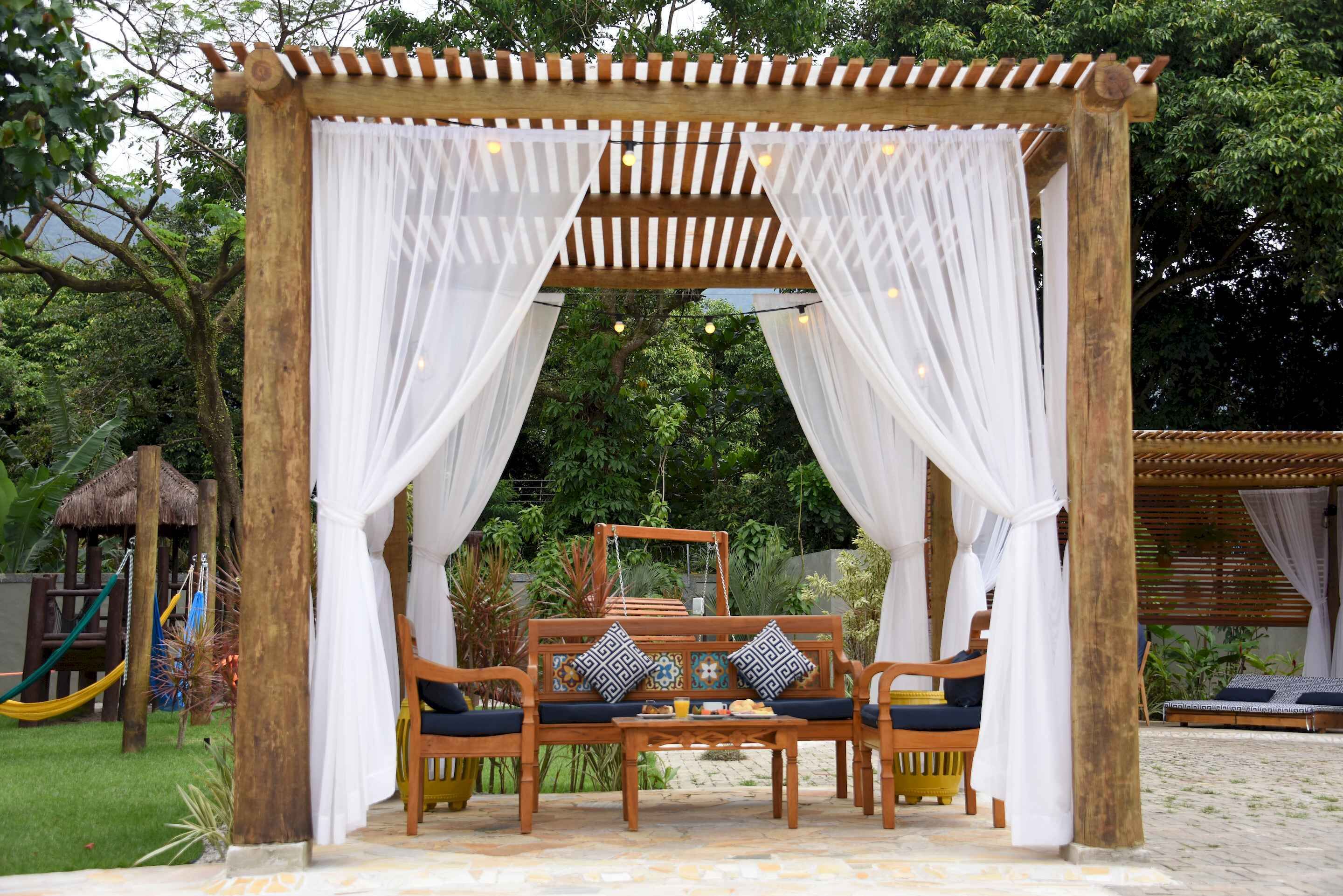 A wooden gazebo with white drapes and a bench with cushions.